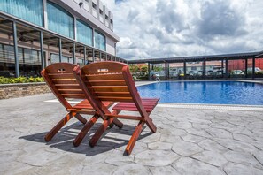 Two wooden lounge chairs by a swimming pool with a modern building in the background.