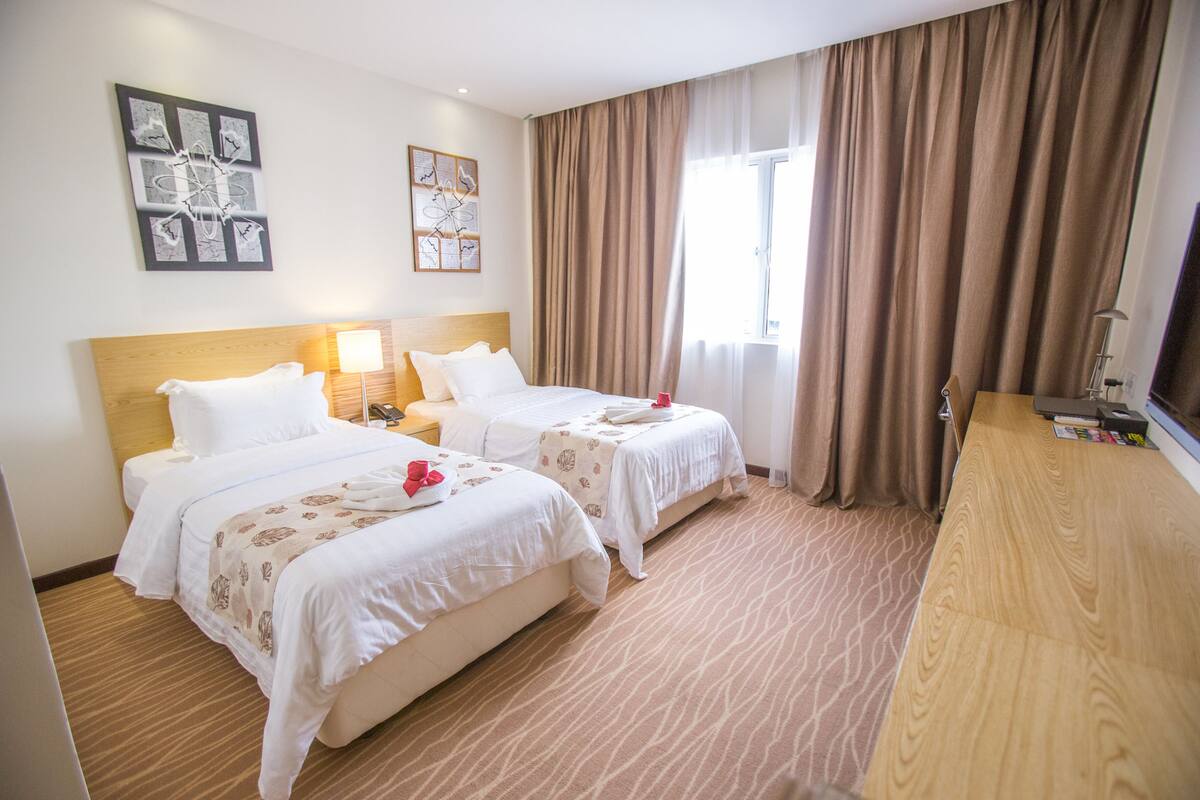 A hotel room with two beds, a wooden headboard, a desk, and a large window with curtains.