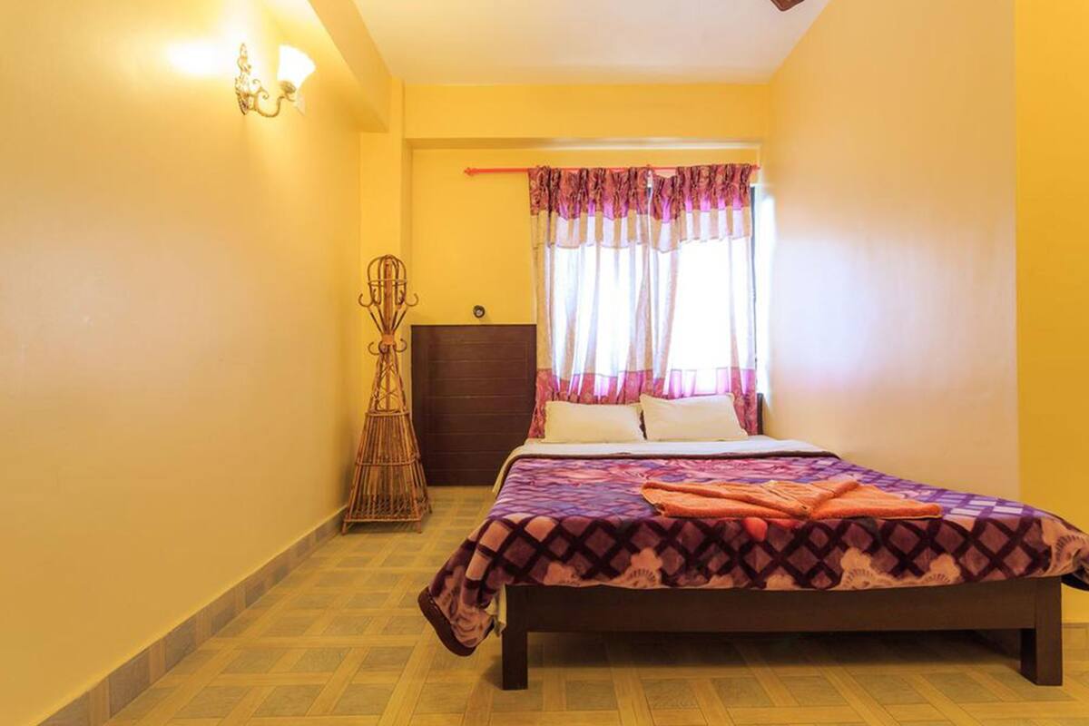 A bedroom with a bed, a bedside table, a lamp, a window with curtains, and a wall-mounted light fixture.