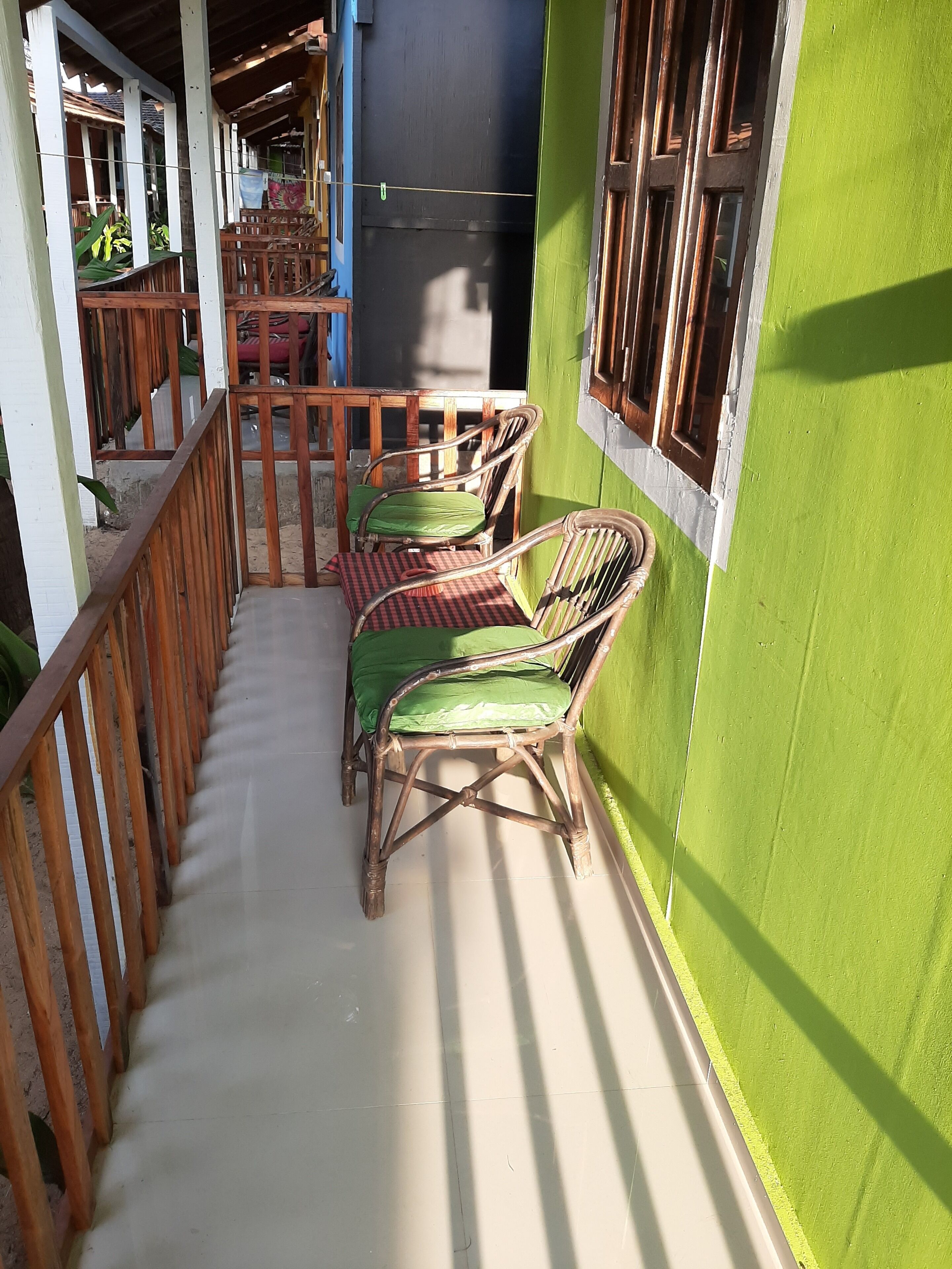 Un balcón con sillas de mimbre, una mesa a cuadros y una baranda de madera, junto a una pared verde.