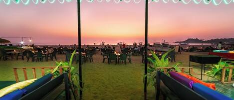A beachside restaurant with outdoor seating and a view of the ocean at sunset.