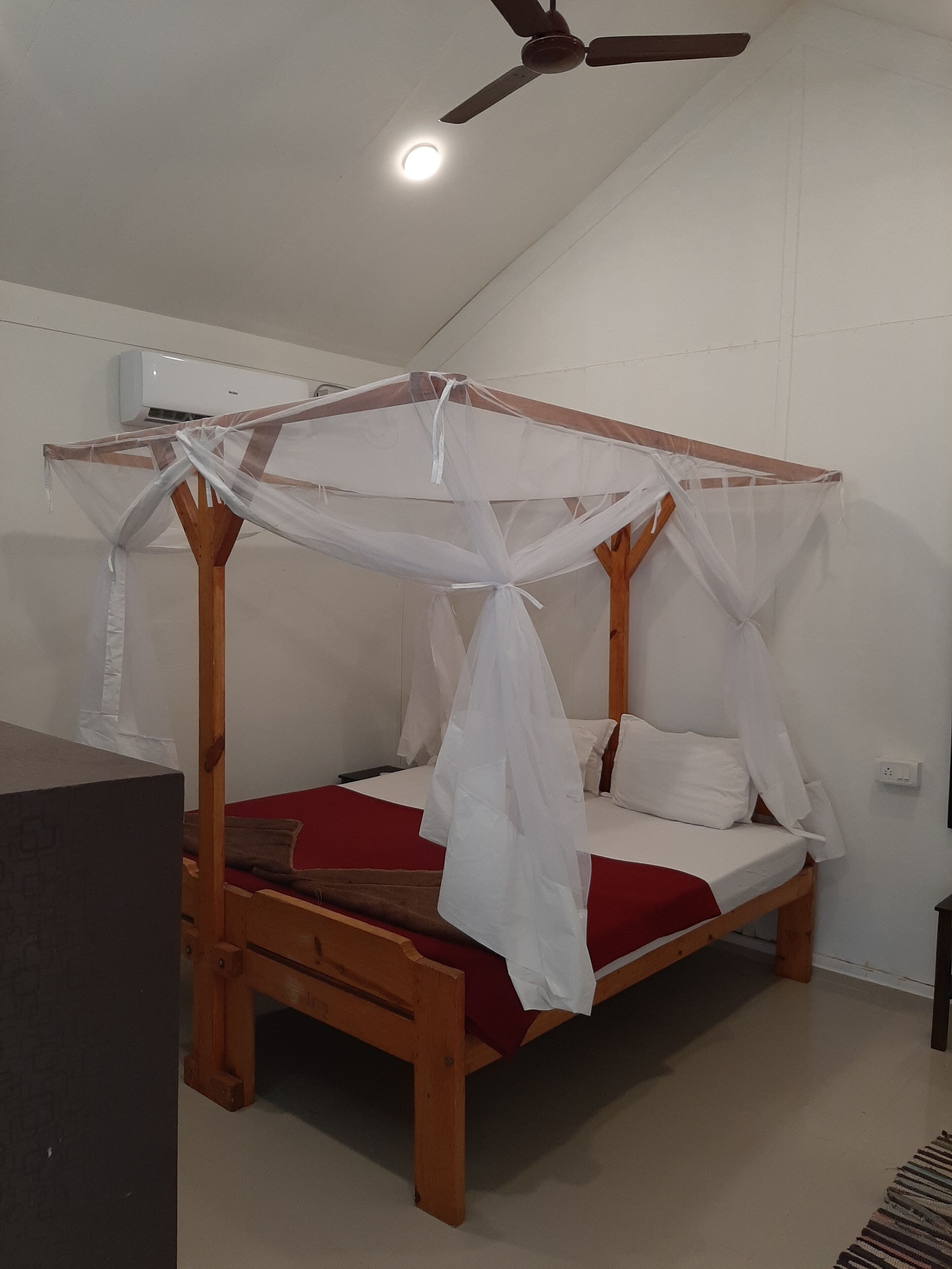 Un lit simple avec un cadre en bois et un ciel de lit blanc, un ventilateur de plafond et un mur blanc sur lequel est installé un appareil de climatisation.