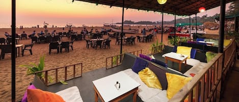 An outdoor seating area with tables and chairs, overlooking a beach with people and boats.