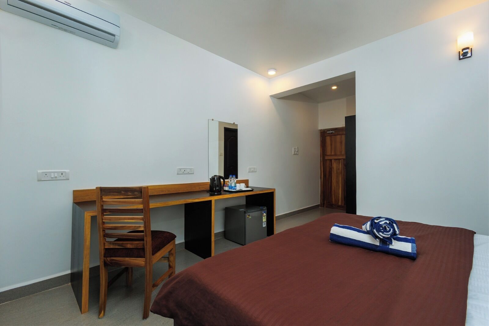 Habitación de hotel pequeña con una cama individual, un escritorio de madera, una silla, un refrigerador pequeño y un espejo.
