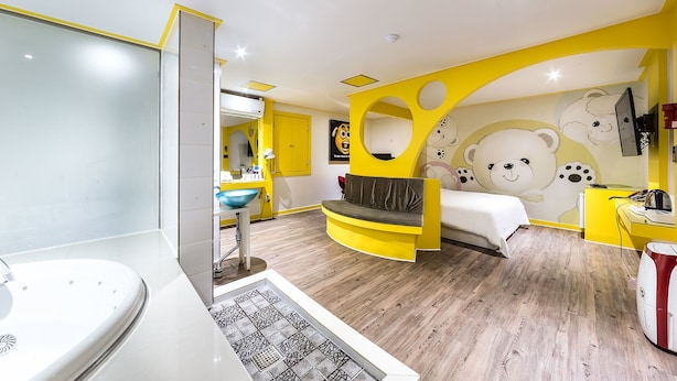 A modern hotel room with a yellow and white color scheme, a large bed, a seating area, a bathroom with a bathtub, and a wall with bear-themed artwork. - In Hotel (Suwon)