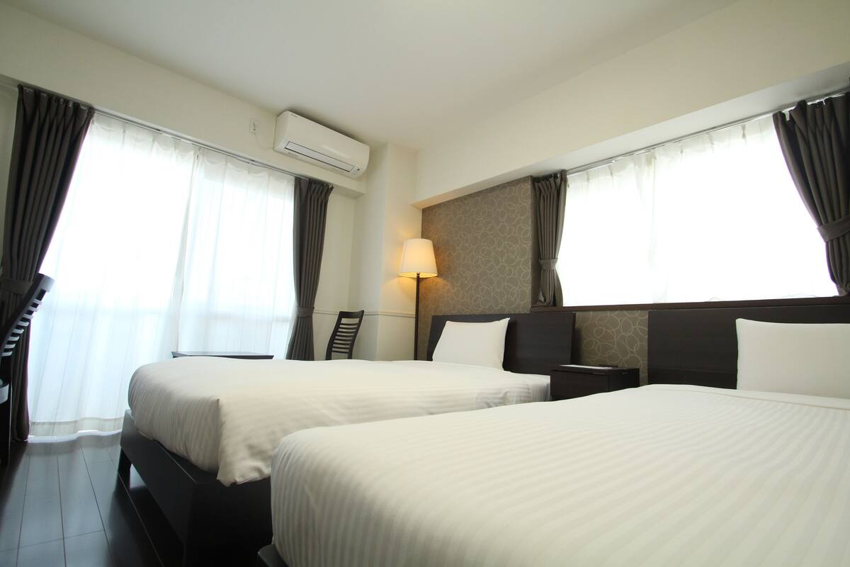 Quarto de hotel com duas camas, uma mesa de cabeceira, um abajur e um aparelho de ar condicionado.