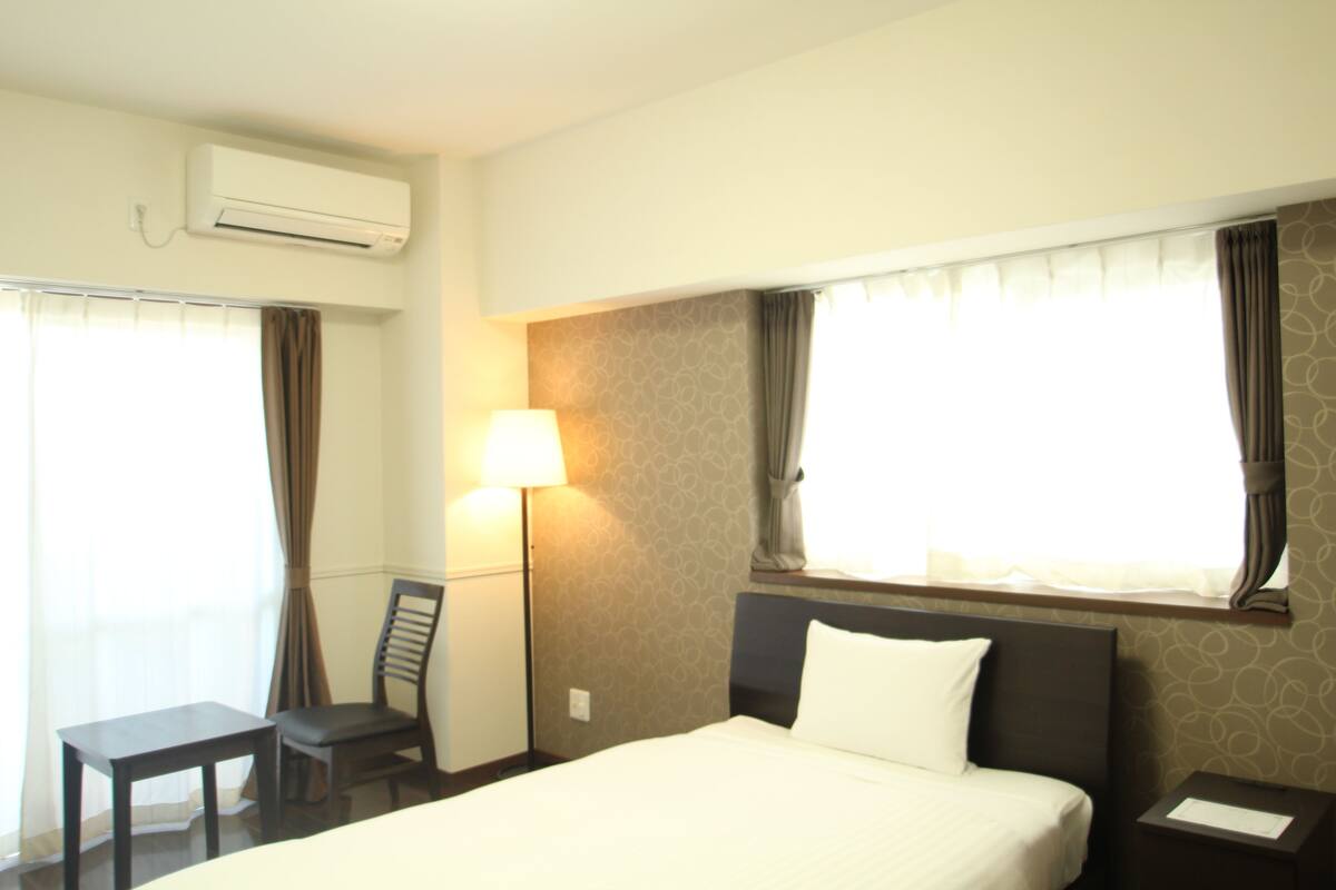 Quarto de hotel com cama, escrivaninha, cadeira, luminária e ar condicionado.