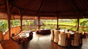 A wooden deck with wicker furniture and a table set for dining, overlooking a lush green landscape. - Dragonfly Village (Ubud)