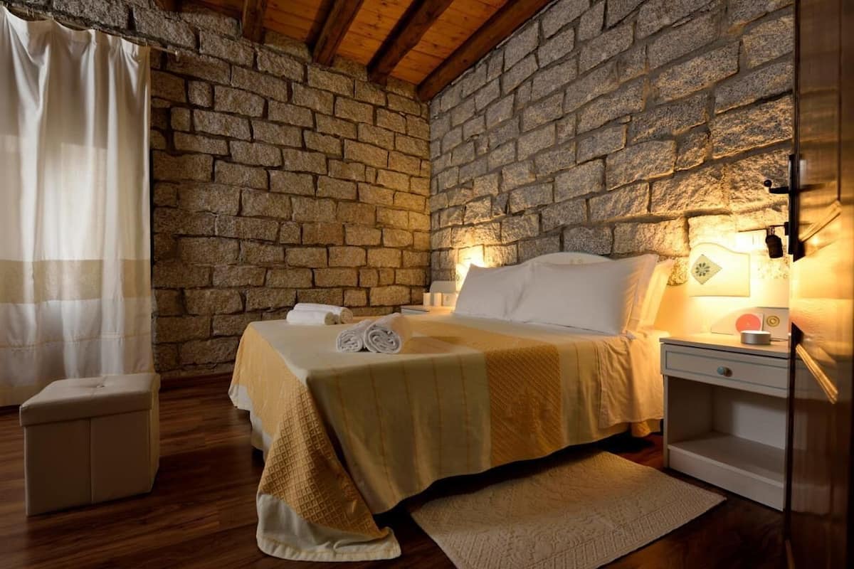 A bedroom with a stone wall, a bed with a yellow blanket, a bedside table with a lamp, and a small bedside table with a radio.