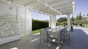 A covered patio area with a table and chairs, a decorative wall, and a view of a garden and trees. - Antigone Estate (Rhodes)