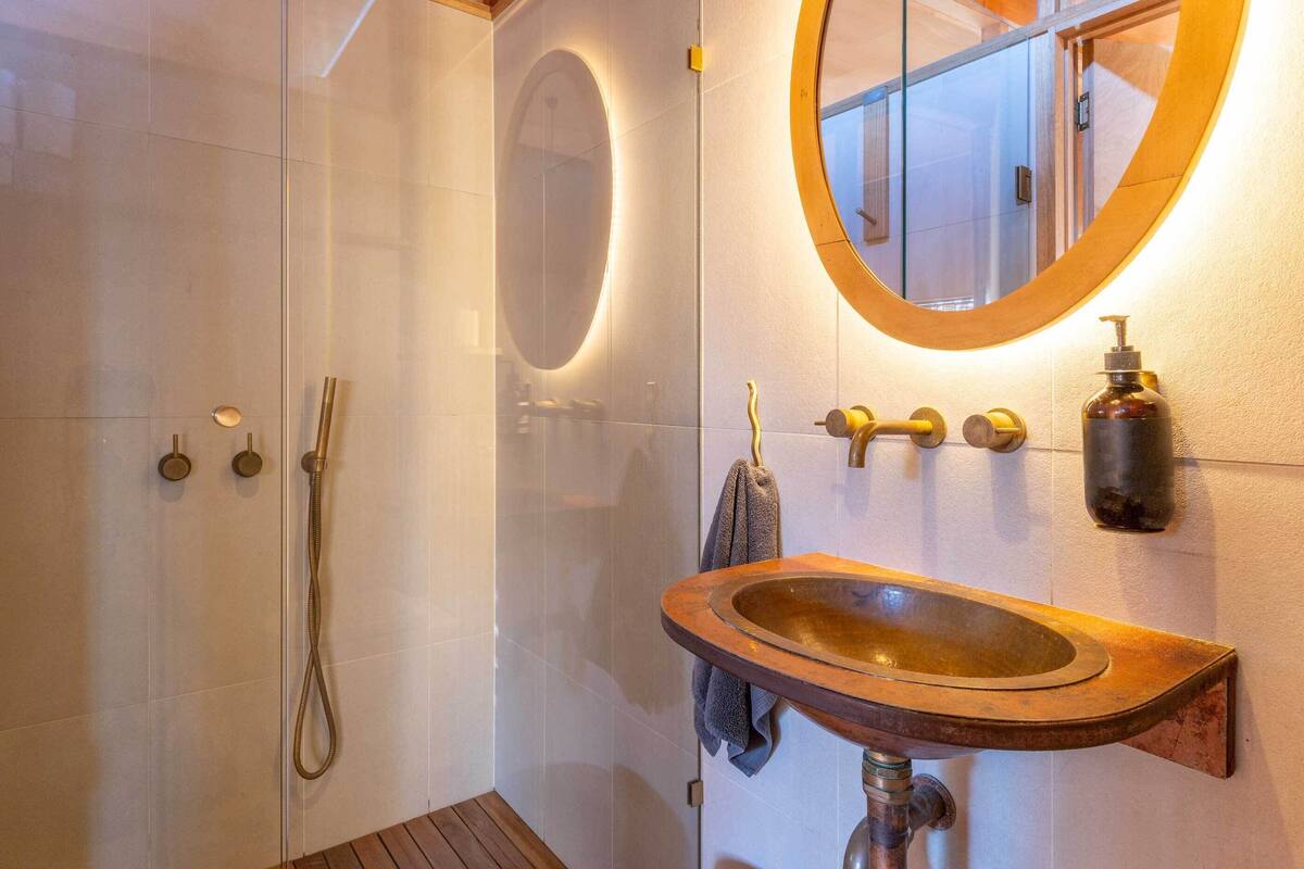 A bathroom with a wooden sink, a round mirror, a handheld showerhead, and a towel hanging on the wall.