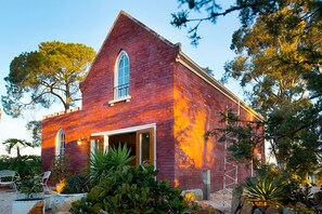 A red brick building with a large arched window and a door, surrounded by various plants and trees.