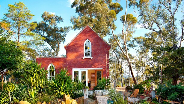 A red brick house with a white door and window, surrounded by a garden with various plants and rocks.