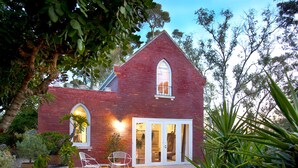 A red brick house with a large arched window and a white door. There are two white chairs outside and a potted plant nearby.