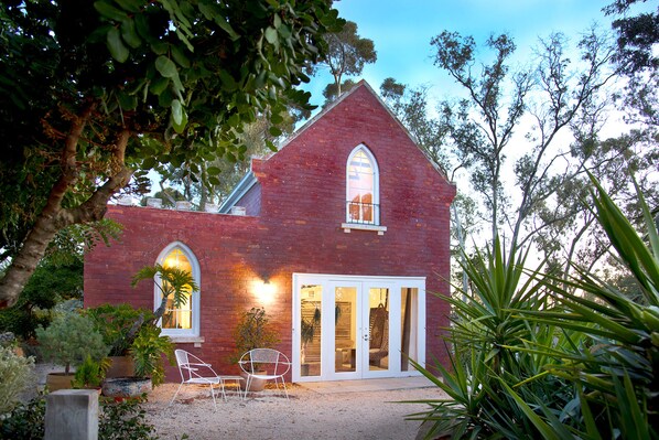 A red brick house with a large arched window and a white door. There are two white chairs outside and a potted plant nearby.