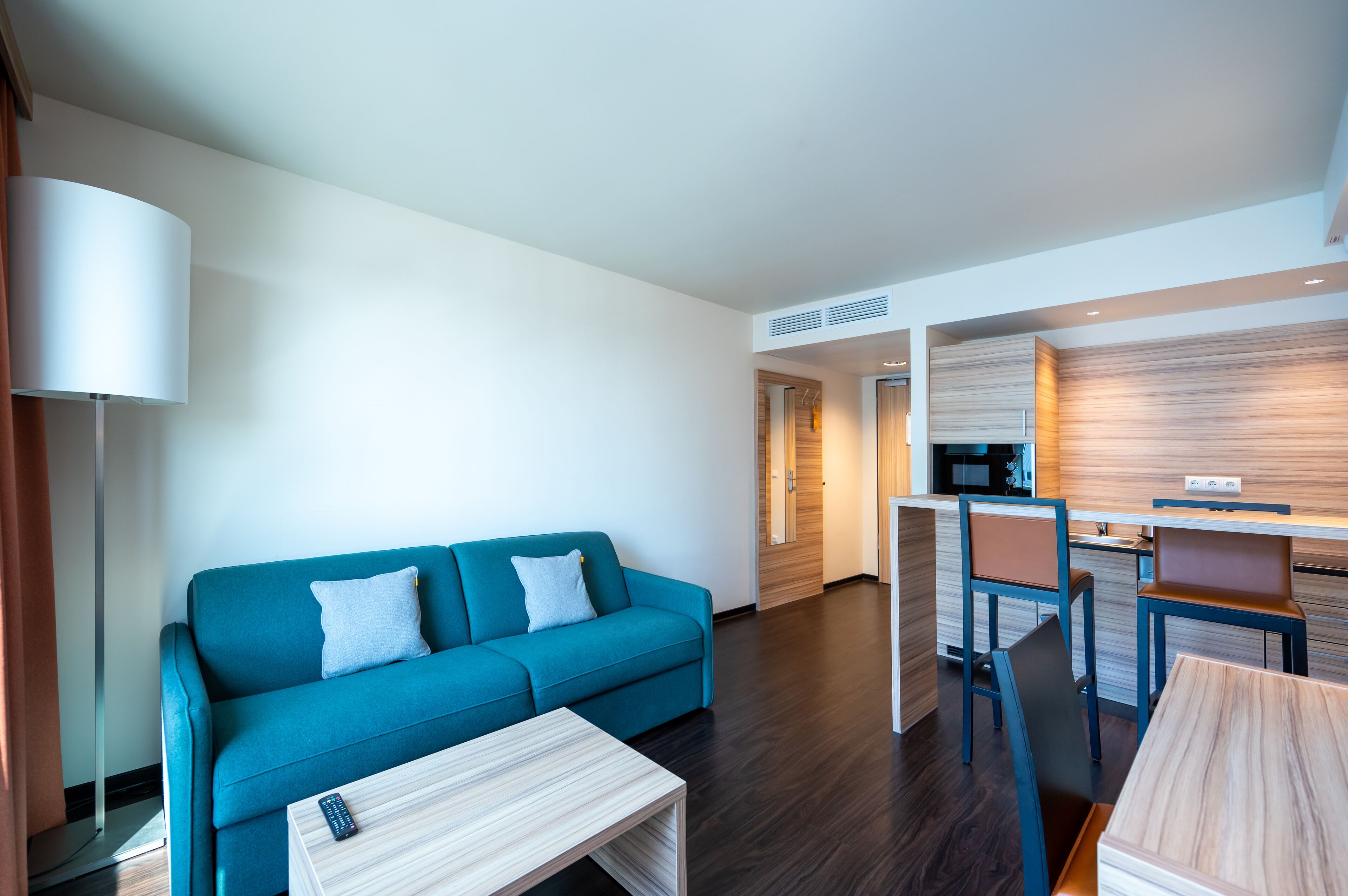 A modern hotel room with a blue sofa, a small wooden table, a desk with a chair, and a kitchen area.