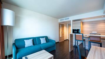 A modern hotel room with a blue sofa, a small wooden table, a desk with a chair, and a kitchen area.