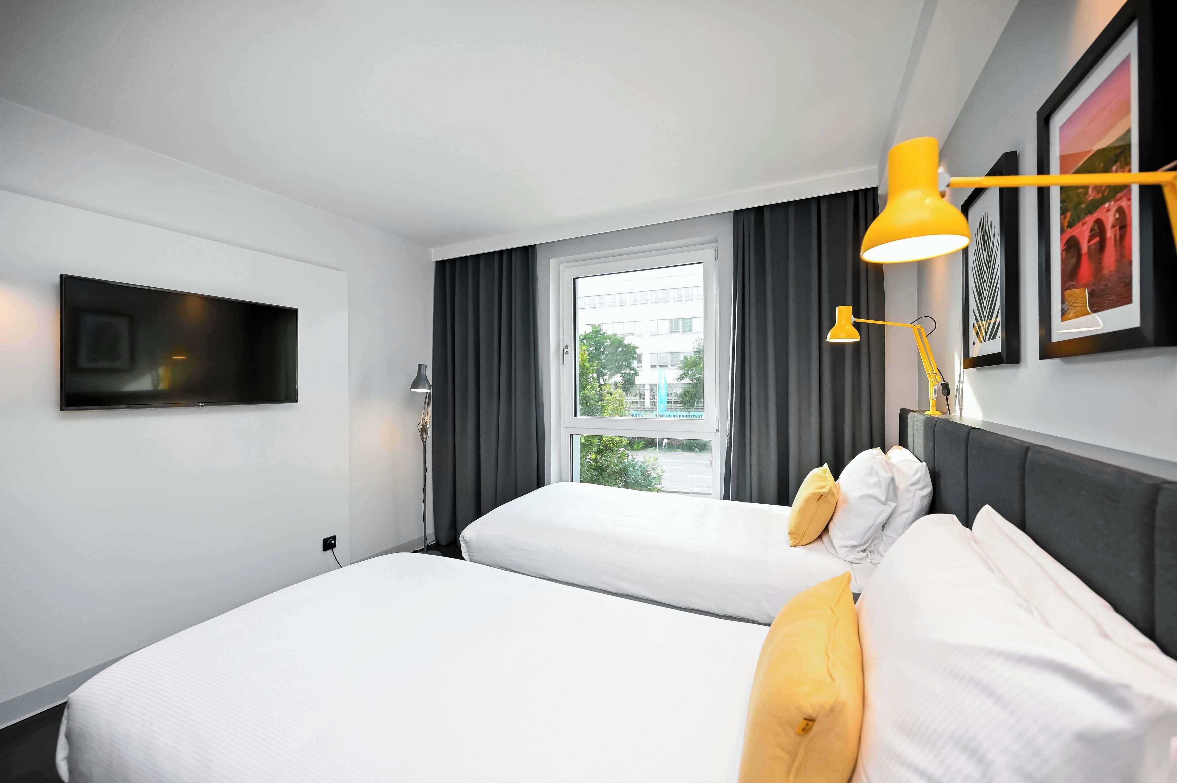 Ein modernes Hotelzimmer mit zwei Betten, einem großen Fenster und einem an der Wand befestigten Flachbildfernseher.