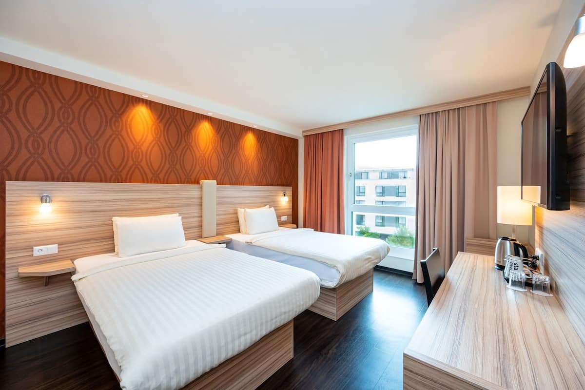 Ein Hotelzimmer mit zwei Betten, einem Schreibtisch, einem Fernseher und einem Fenster mit Vorhängen.