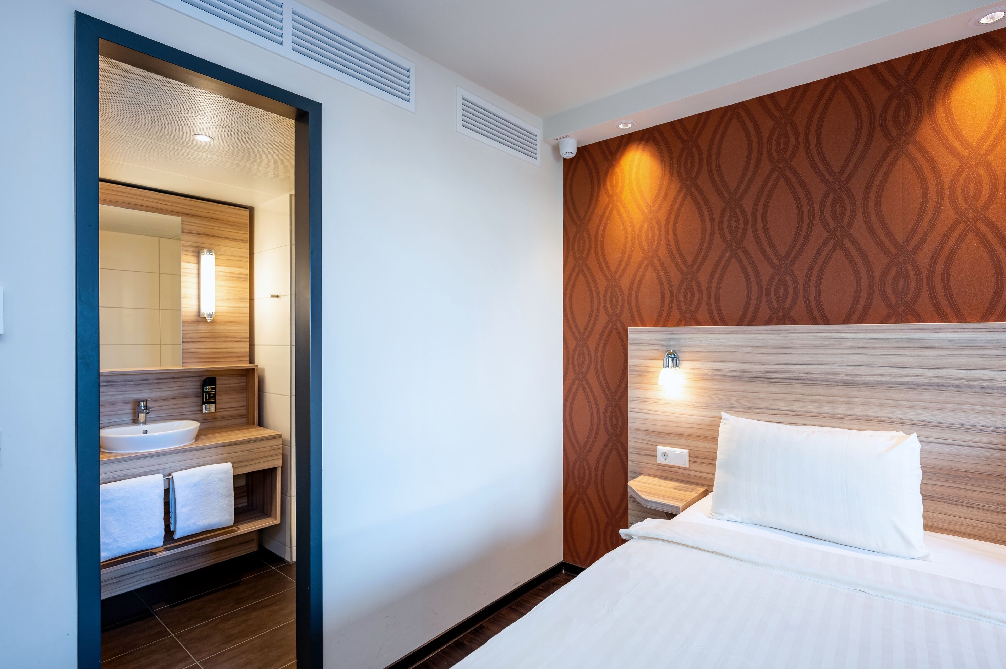 A modern hotel room with a wooden headboard, a large mirror, a built-in sink, and a neatly made bed.