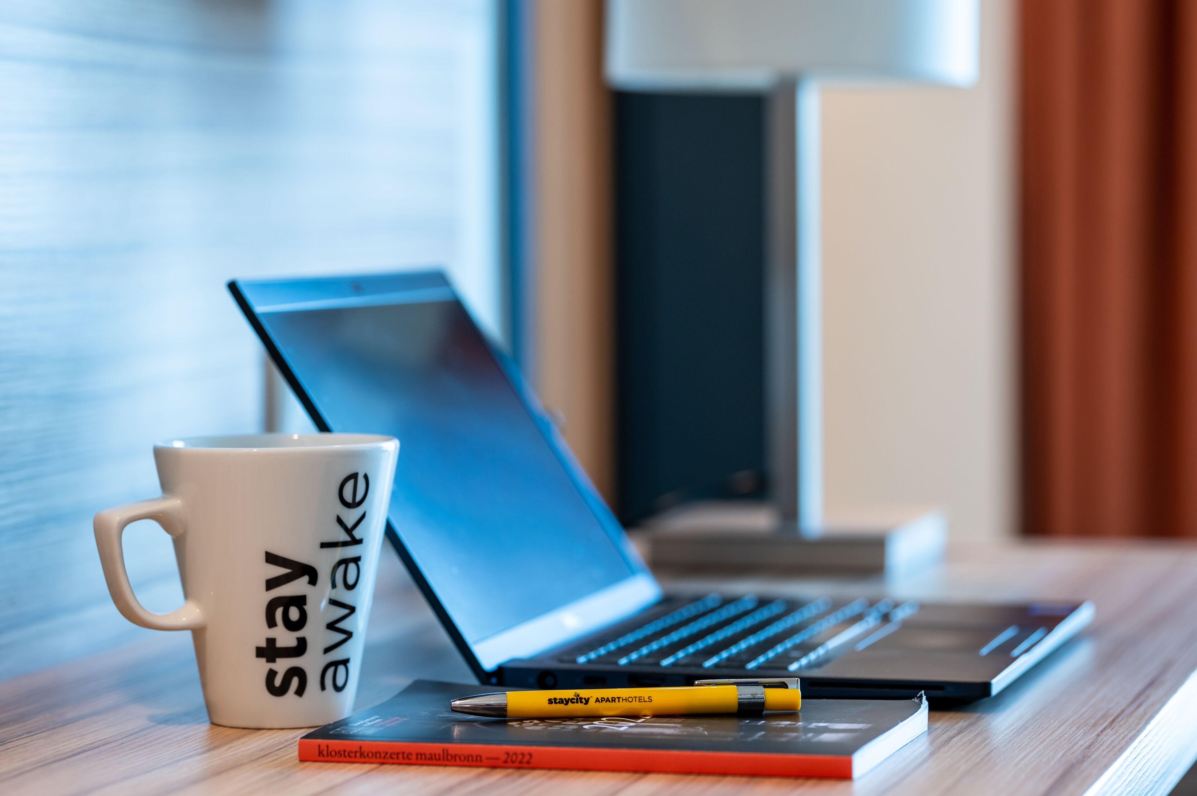 Un ordinateur portable, une tasse blanche avec l’inscription « restez éveillé », un stylo jaune et un livre rouge sur une table en bois.