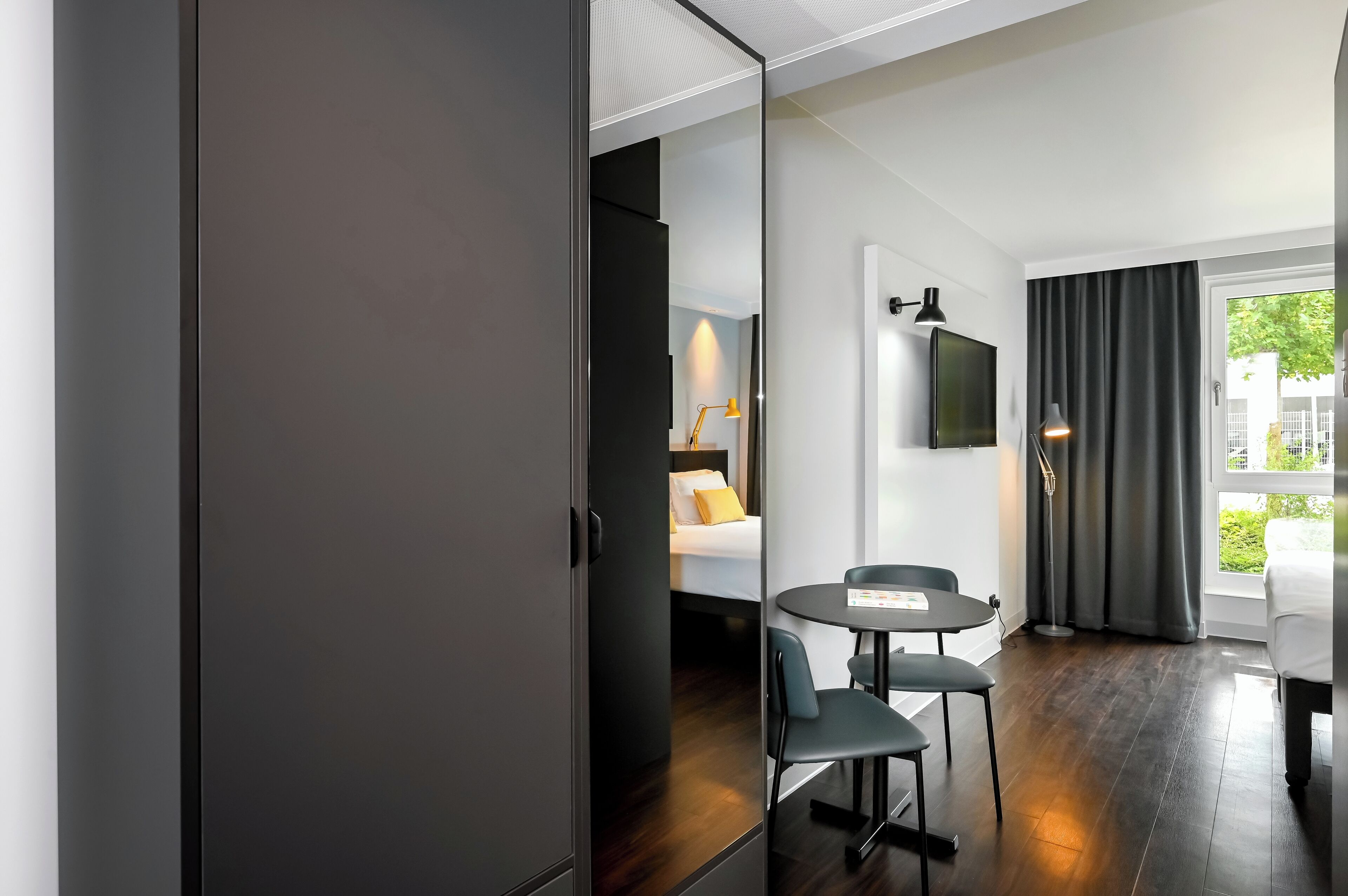 A modern hotel room with a bed, a desk with two chairs, a flat-screen TV, and a large window with curtains.