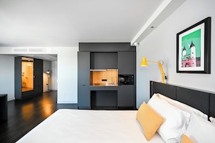 Quarto de hotel moderno com uma cama grande, escrivaninha embutida e uma pequena cozinha equipada.