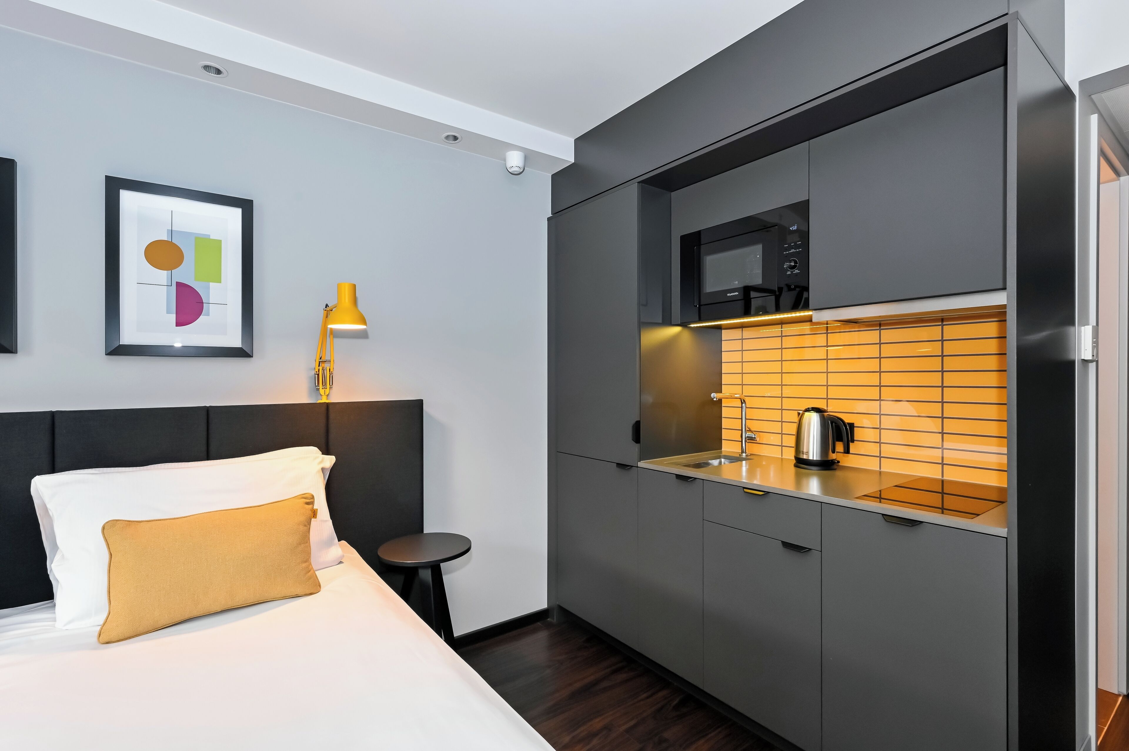 Habitación de hotel compacta con cama, mesita de noche y kitchenette equipada con microondas y tetera.