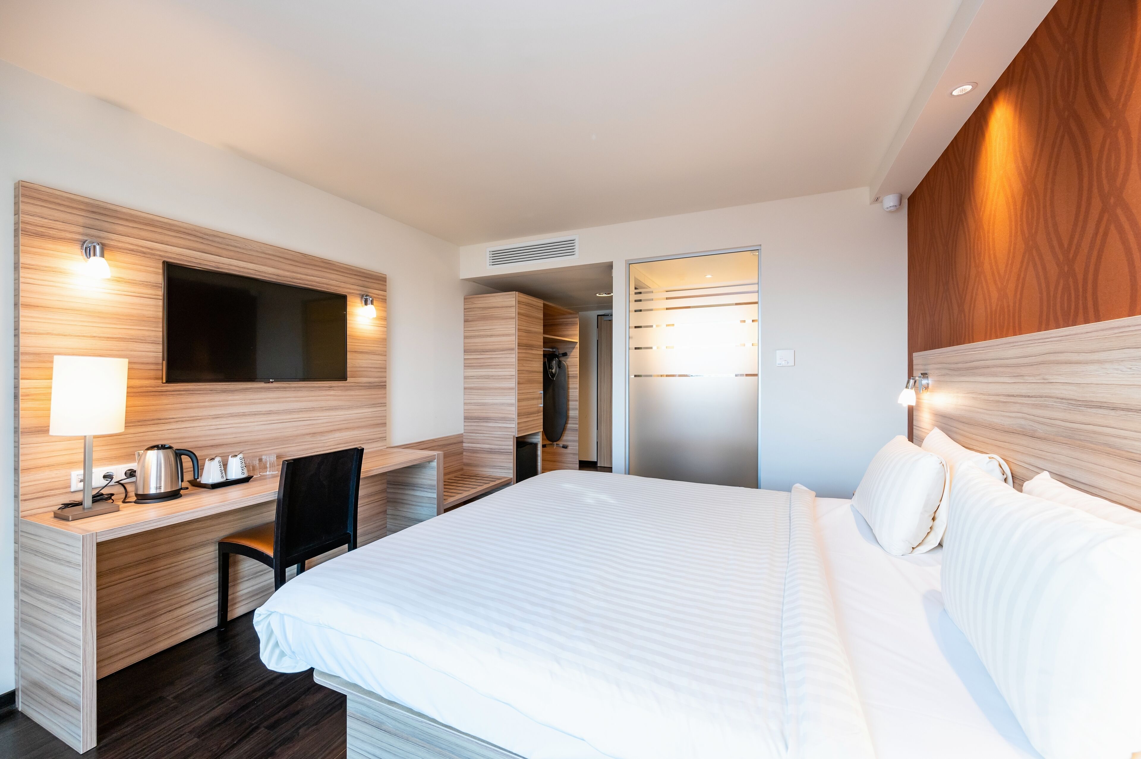 A modern hotel room with a large bed, a desk with a kettle, a chair, and a TV mounted on the wall.