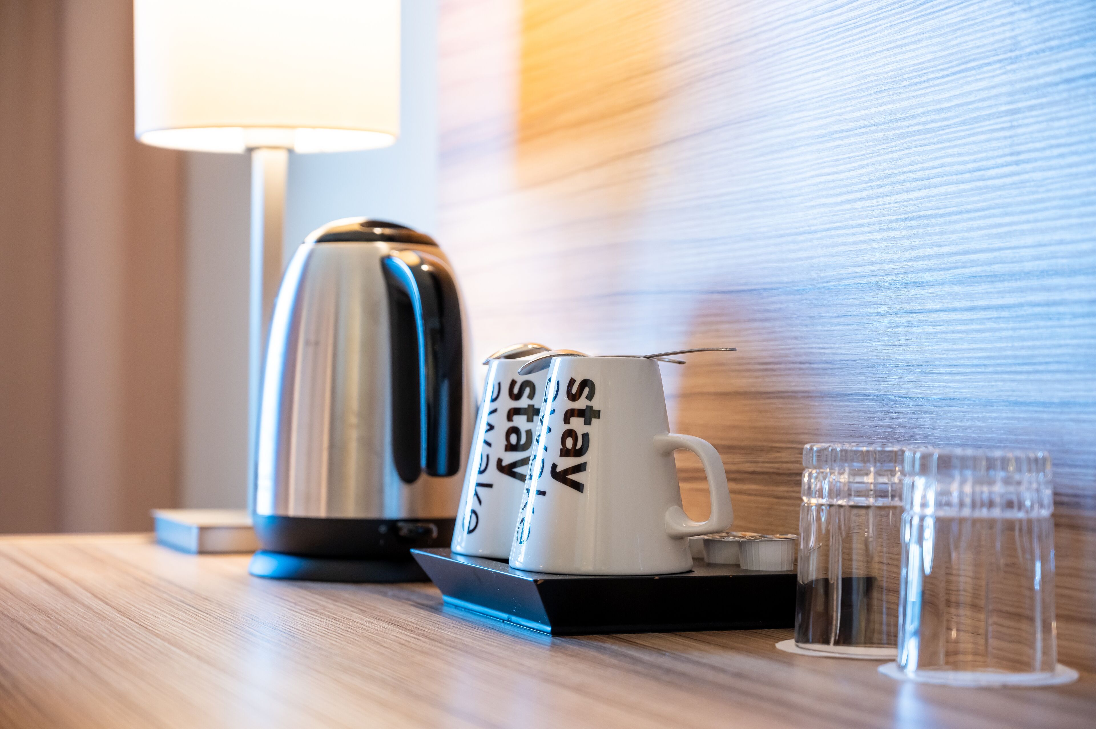 Une chambre d’hôtel équipée d’une machine à café, de deux tasses sur lesquelles est inscrit le mot « séjour », et de deux verres disposés sur un plateau.