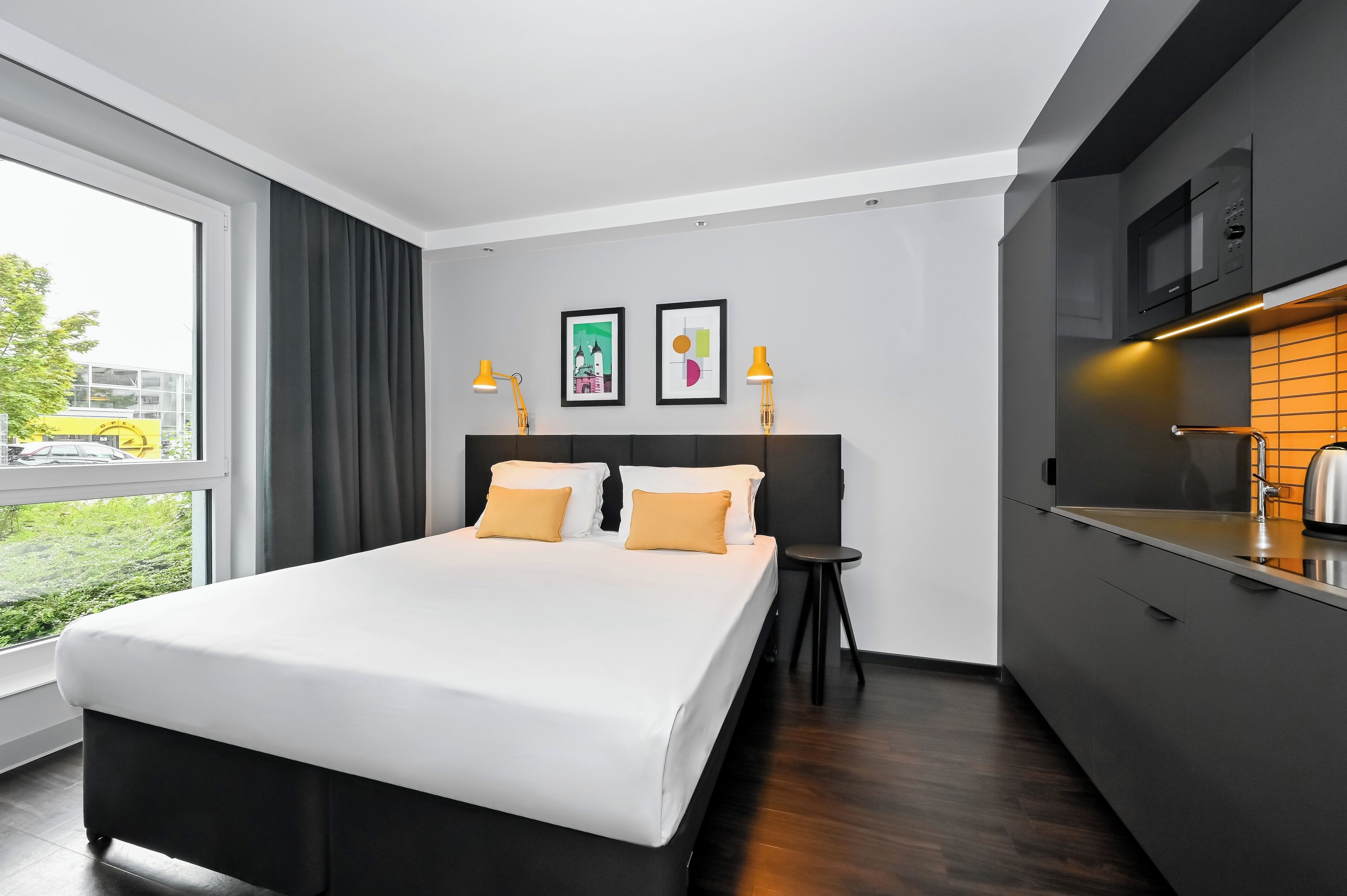 A modern hotel room with a large bed, a small kitchen area, and a view of the outdoors.