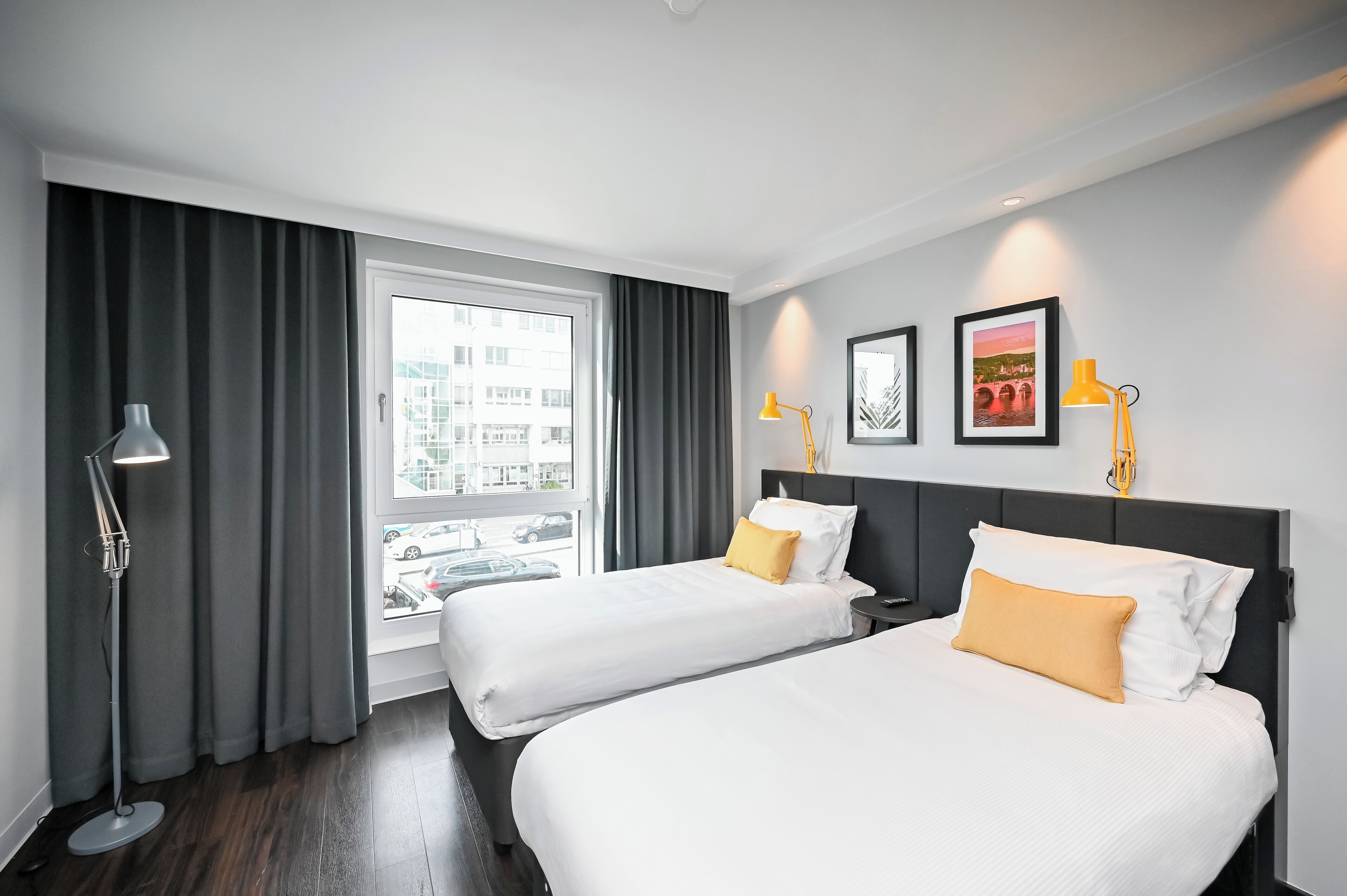 A hotel room with two beds, a large window with curtains, a floor lamp, and two wall-mounted lamps.