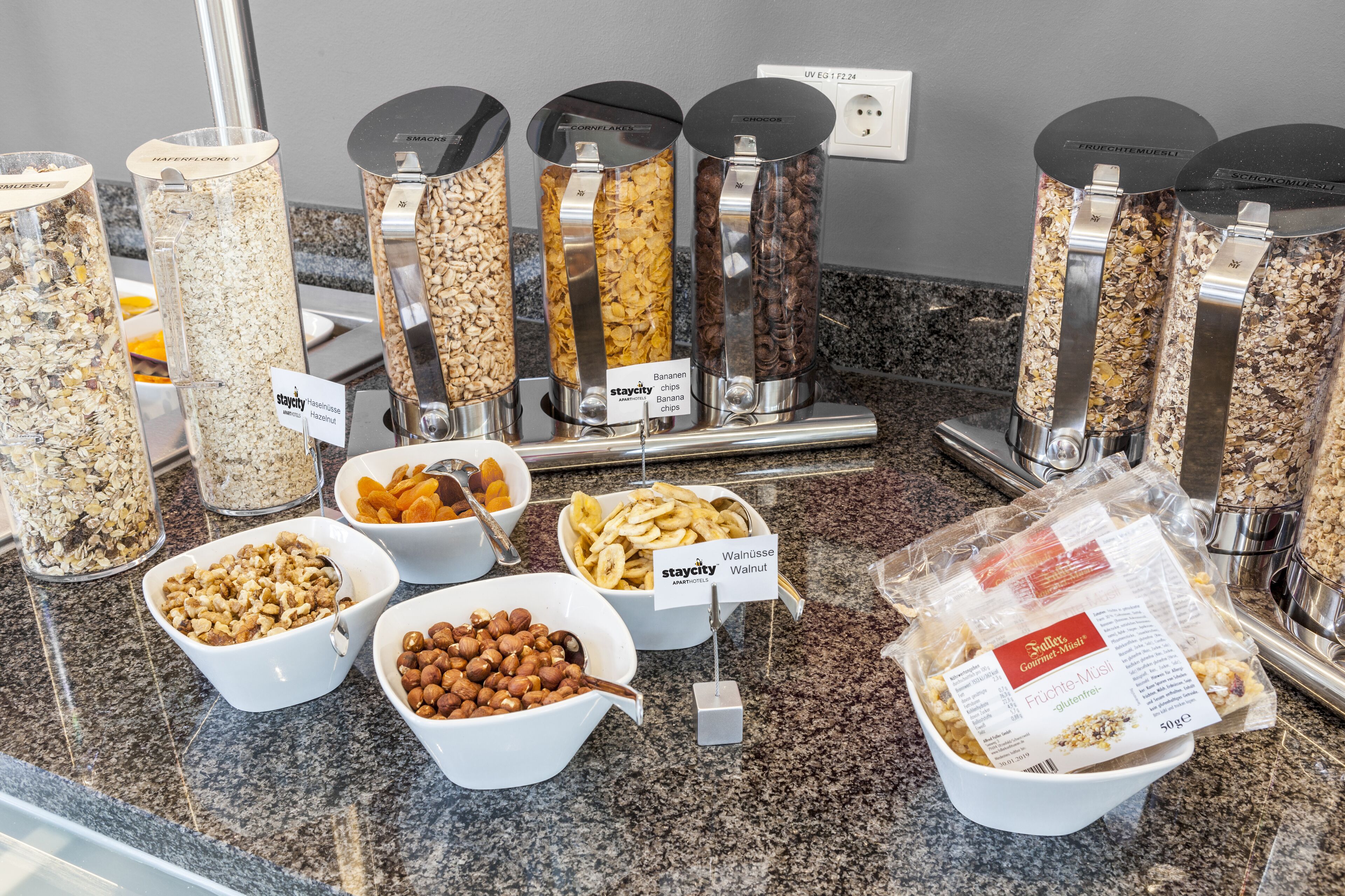 A variety of food items including granola, nuts, banana chips, and chocolate chips displayed in bowls and containers.
