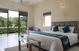 A bedroom with a large bed, a glass table, and a view of greenery outside.