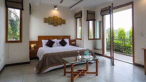 Ein Hotelzimmer mit einem großen Bett, einem Glastisch, einem Deckenventilator und einem Balkon mit Blick auf Grünflächen.