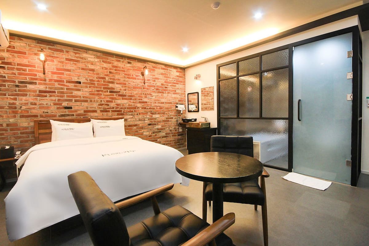 Habitación de hotel con pared de ladrillo, cama con ropa blanca, una mesa redonda, dos sillas y una puerta corrediza de vidrio.