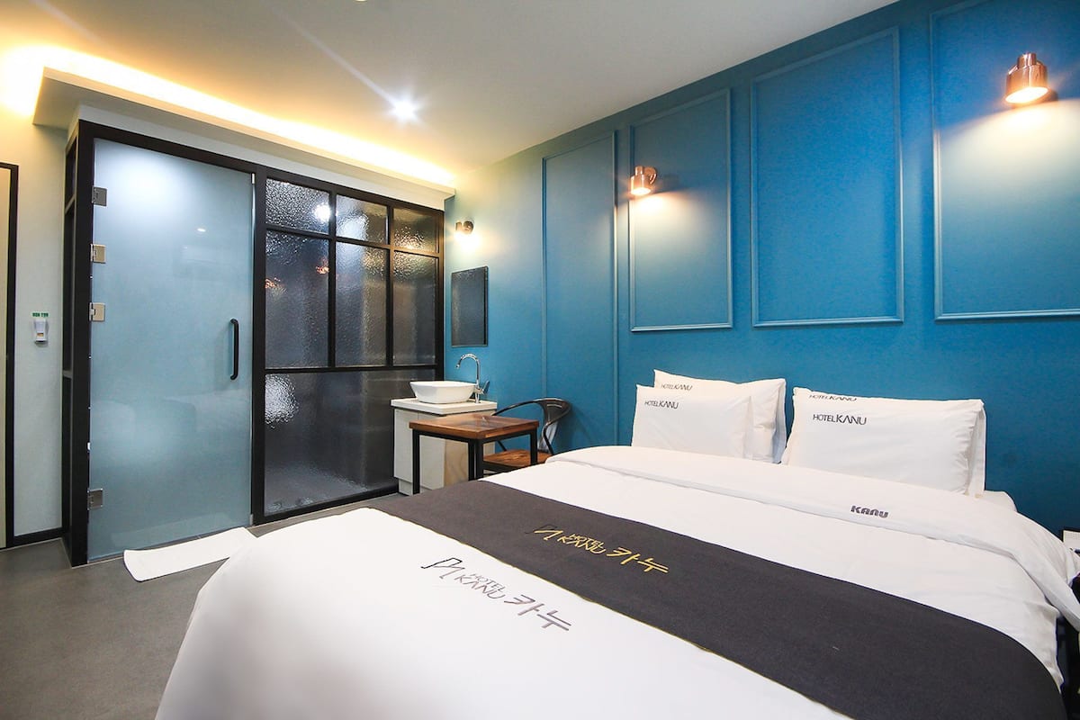 Una habitación de hotel con una cama, una mesita de noche, un espejo, una ventana con cristal esmerilado y una puerta con un panel de cristal satinado.