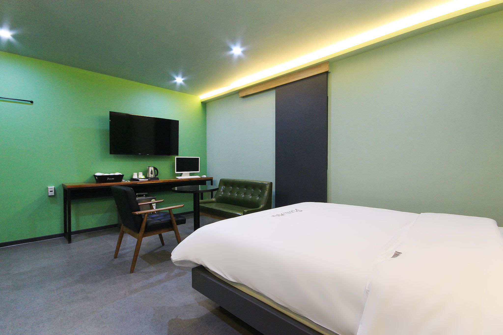 Habitación de hotel con cama, escritorio con computadora, silla, televisión montada en la pared y ventana con persiana oscura.