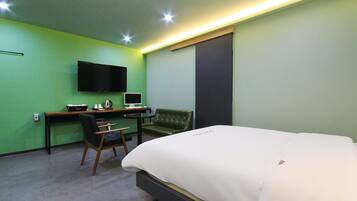 Habitación de hotel con cama, escritorio con computadora, silla, televisión montada en la pared y ventana con persiana oscura.
