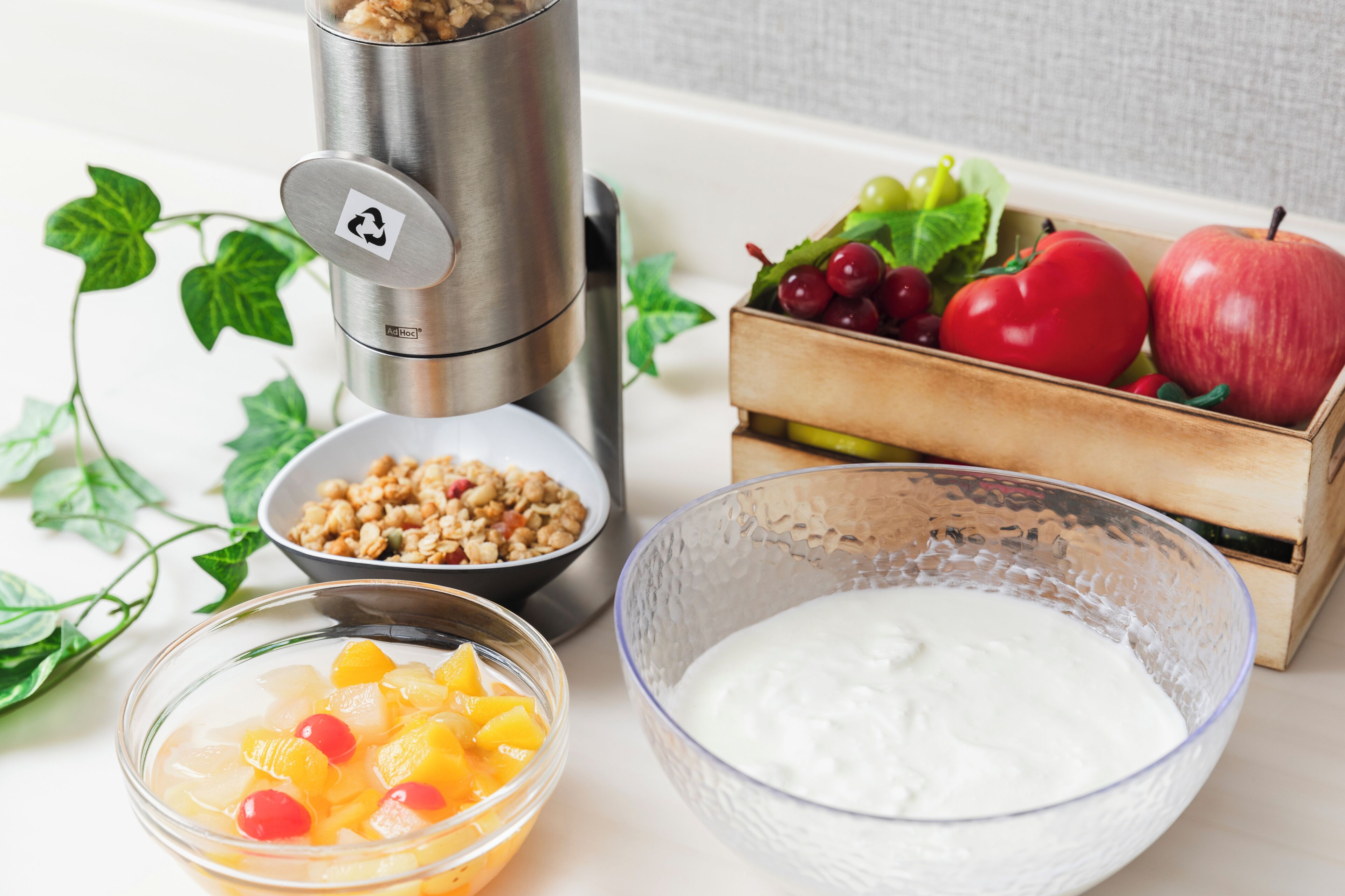 A kitchen counter with a stainless steel coffee maker, a bowl of granola, a bowl of fruit salad, a bowl of yogurt, and a wooden crate containing apples and grapes.