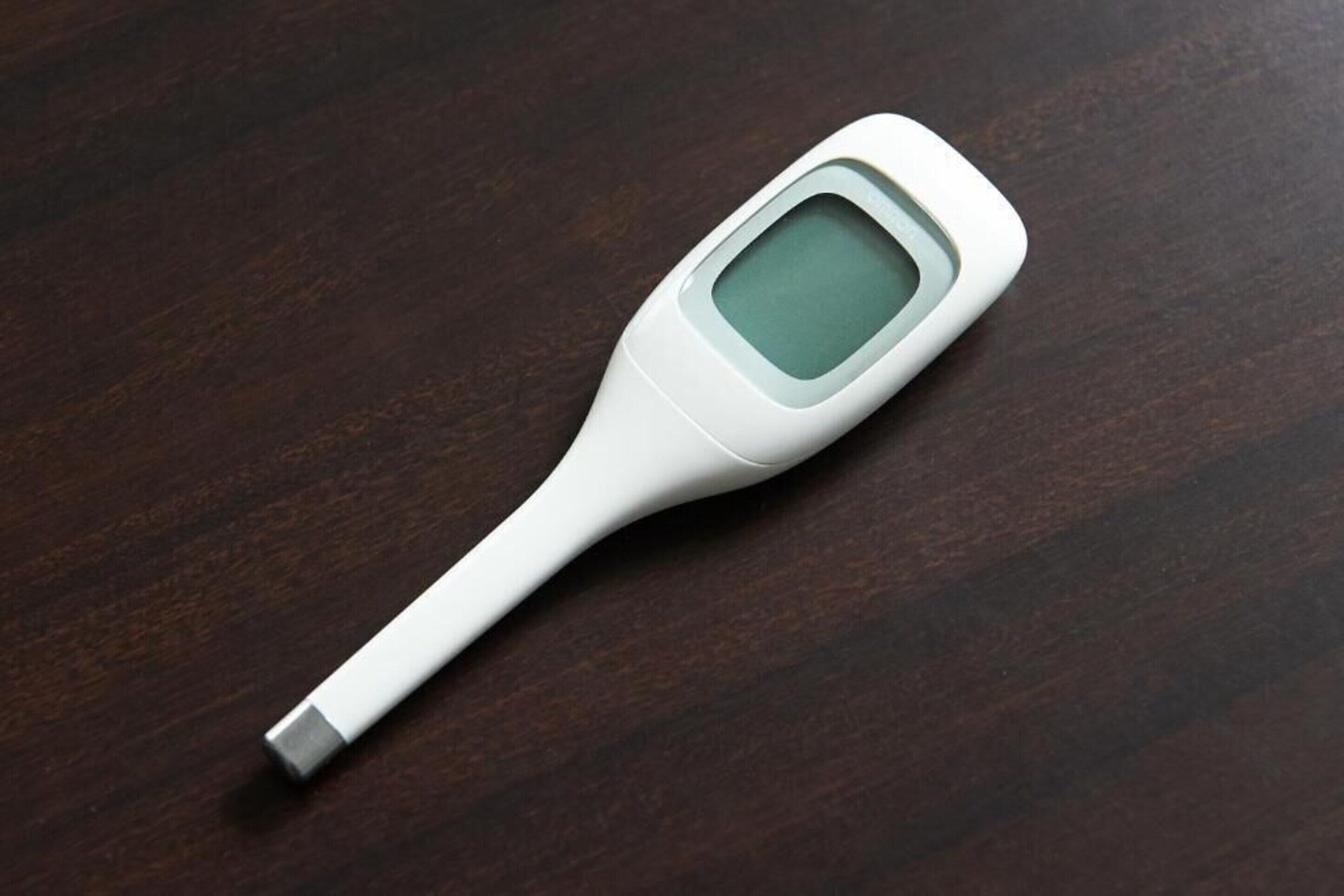 A digital thermometer with a white body and a metallic tip, placed on a dark wooden surface.