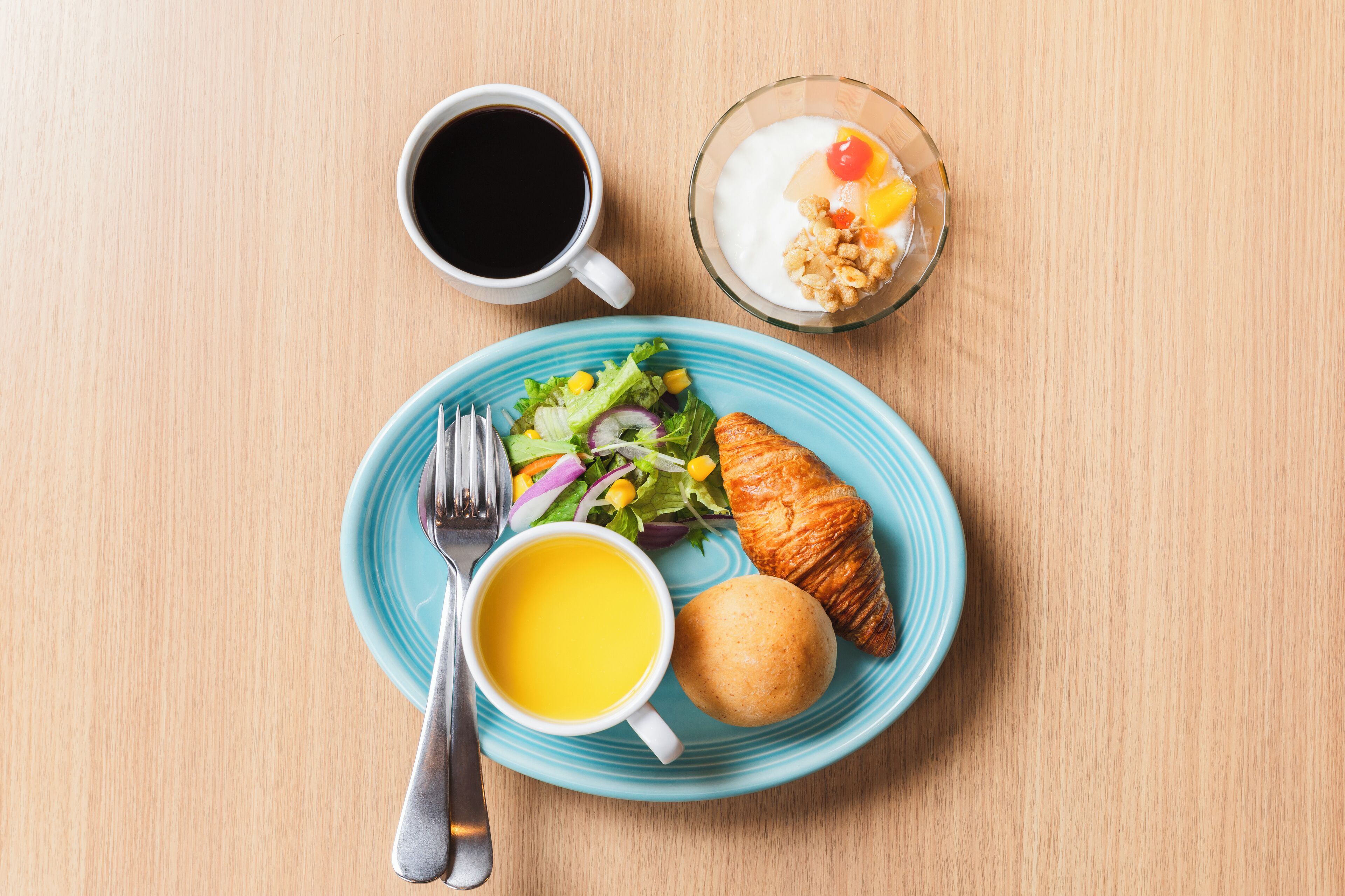 Une table dressée pour le petit-déjeuner avec un croissant, un bol de yaourt aux fruits, une tasse de café et une assiette de salade.