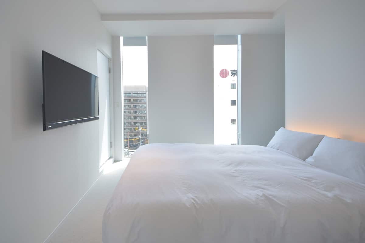 Une chambre moderne avec un grand lit, un téléviseur à écran plat fixé au mur et une vue sur le paysage urbain, visible à travers une grande fenêtre.
