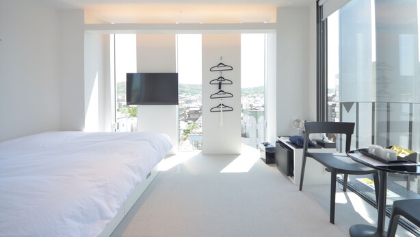 Quarto de hotel moderno com uma cama grande, uma escrivaninha com cadeira, televisão e vista para a cidade através de amplas janelas.