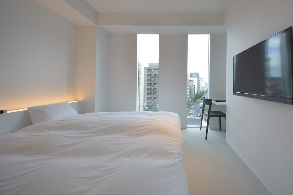 Une chambre d’hôtel moderne, dotée d’un grand lit, d’un bureau et d’une télévision. De grandes fenêtres offrent une vue sur la ville.
