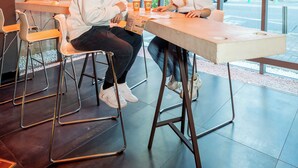 Duas pessoas sentadas à mesa em um espaço interno moderno, com grandes janelas e luminária pendente com design curvo.