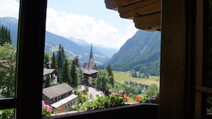 Vista desde una ventana con vista a un pueblo de montaña con una iglesia, casas y abundante vegetación.