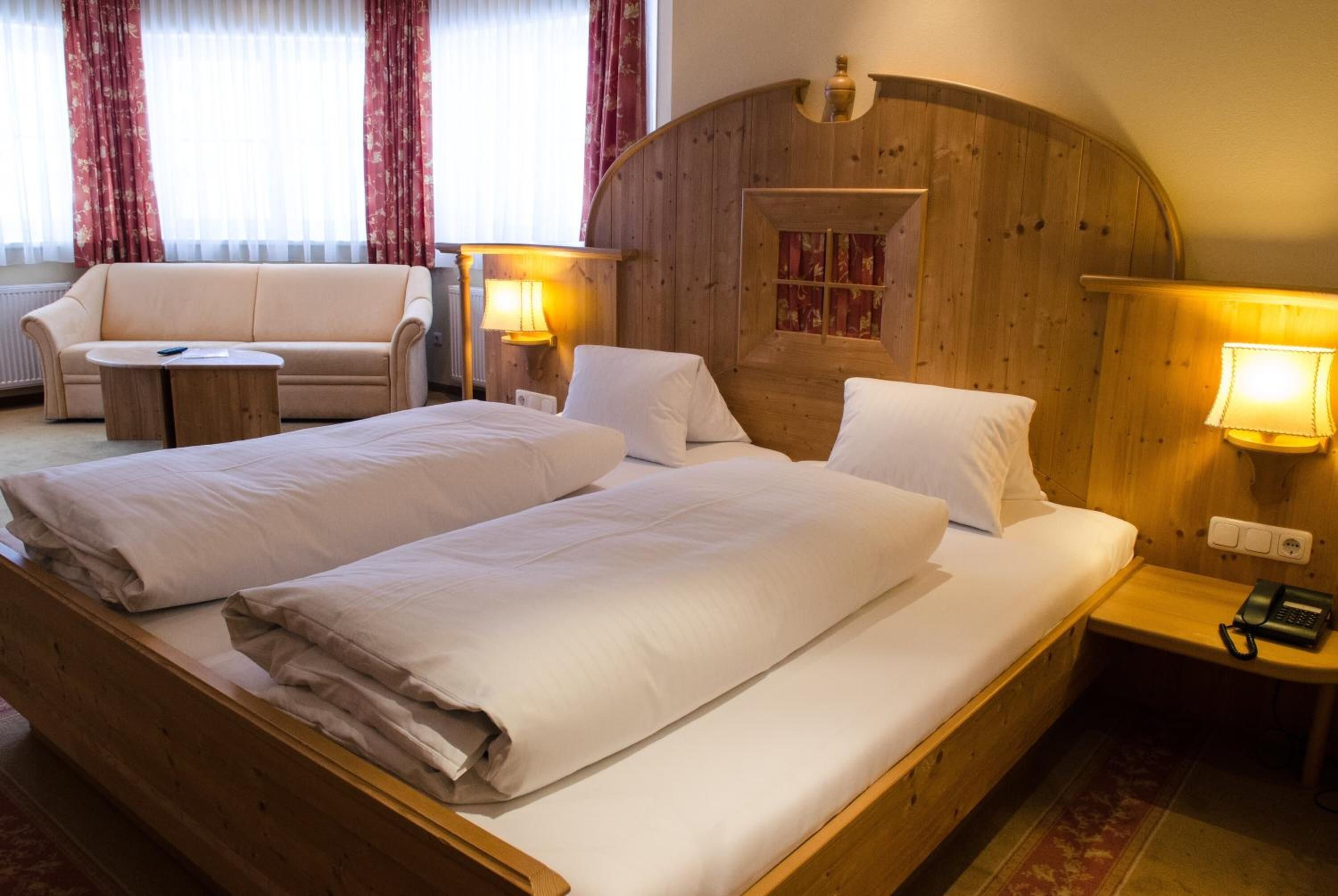 A double bed with white linens, a wooden headboard, and two bedside lamps. There is a wooden nightstand with a phone and a small table with a lamp. A beige sofa and a small round table are also visible.