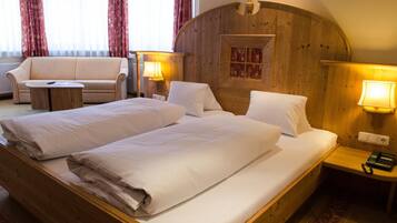 A double bed with white linens, a wooden headboard, and two bedside lamps. There is a wooden nightstand with a phone and a small table with a lamp. A beige sofa and a small round table are also visible.