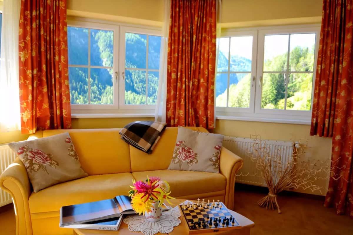 Ein gelbes Sofa mit floralen Kissen, ein Schachbrett und ein Buch mit dem Titel "KARNTEN" auf einem Tisch.